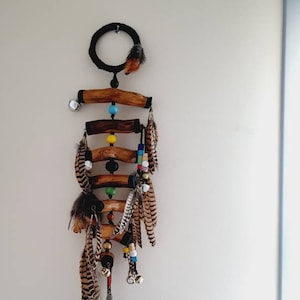 Puede incluir: Un atrapasueños de madera con plumas, cuentas y campanas. El atrapasueños está hecho de madera natural y tiene una parte superior circular con un cordón negro. Las plumas son marrones y blancas, y las cuentas son de colores.