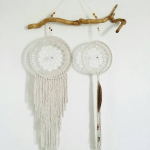 Op de afbeelding: Twee witte macramé droomvangers die aan een houten tak hangen. De grotere droomvanger heeft een lange franje en de kleinere droomvanger heeft een enkele bruine veer en kralen.