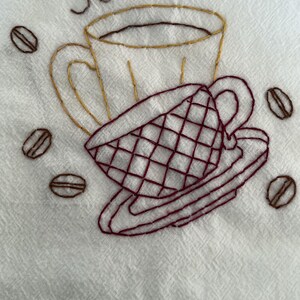 May include: White kitchen towel with embroidered coffee-themed design. Features the word "Java" in brown script above two coffee cups and coffee beans. The cups are in brown and gold thread, with a cross-hatched pattern on the lower cup.