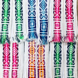 Puede incluir: Diez tiras de tejido blanco tejido con patrones geométricos coloridos. Las tiras tienen flecos en la parte inferior y están dispuestas en dos filas.