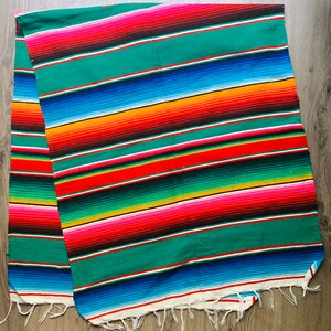 Könnte beinhalten: Eine farbenfrohe Serape-Decke mit Streifen in Grün, Rot, Orange, Blau und Gelb. Die Decke hat einen weißen Fransenbesatz an den Kanten und besteht aus gewebtem Stoff. Die Decke ist etwa 180 cm lang.
