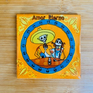 May include: A yellow and orange ceramic tile with a blue circle border. The circle contains a cartoon image of two skeletons, a male and a female, with the text "Amor Eterno" above them.