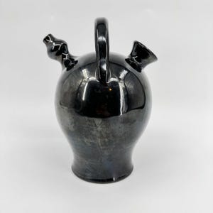 May include: A black ceramic pitcher with a glossy finish. The pitcher has a rounded body, a handle, and two spouts. The surface reflects light, creating a metallic sheen. The design is simple and elegant, suitable for serving beverages.