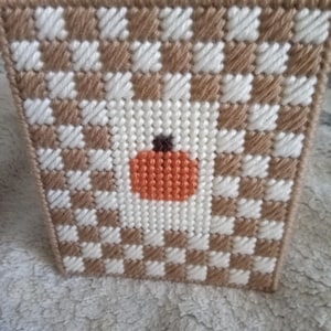 May include: A square tissue box cover crafted from plastic canvas with a woven checkerboard pattern in tan and white yarn. The center of the front panel features a small, stitched orange pumpkin with a dark brown stem on a white background.
