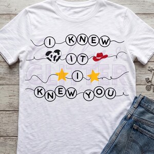 Pu&ograve; includere: Una t-shirt bianca a maniche corte con un design grafico che riporta il testo I KNEW IT I KNEW YOU. Il design include lettere nere all'interno di cerchi, un cuore con stampa mucca bianco e nero, un cappello da cowboy rosso e due stelle gialle.