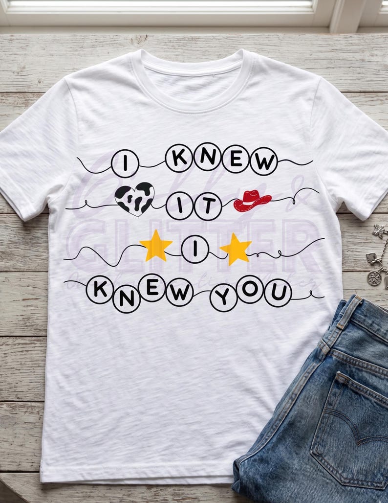 Pu&ograve; includere: Una t-shirt bianca a maniche corte con un design grafico che riporta il testo I KNEW IT I KNEW YOU. Il design include lettere nere all'interno di cerchi, un cuore con stampa mucca bianco e nero, un cappello da cowboy rosso e due stelle gialle.