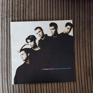 Può includere: Una copertina di un CD con una foto di cinque uomini vestiti con abiti scuri su sfondo bianco. Il testo "boyzone said and done tour" è stampato in bianco. Gli uomini guardano la fotocamera, con un uomo che appoggia il braccio sulla testa.