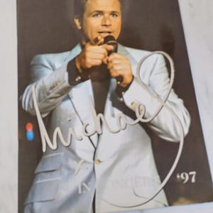 Puede incluir: Un programa de concierto rectangular con una fotografía de un artista con traje azul claro y camisa negra sosteniendo un micrófono. La portada muestra el texto cursivo blanco Michael, la palabra IN CONCERT y el año 97 en fuente blanca.