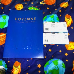 Pode incluir: Uma pasta azul com o texto dourado "BOYZONE THANK YOU & GOODNIGHT" é exibida ao lado de dois bilhetes brancos. Os itens estão em um tecido azul com um padrão de planetas e estrelas.