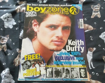 May include: A Boyzone magazine featuring Keith Duffy on the cover. The magazine has a blue and yellow color scheme with the text "100% Official!" and "Exclusive new interview inside!" The background is a black fabric with dog illustrations.