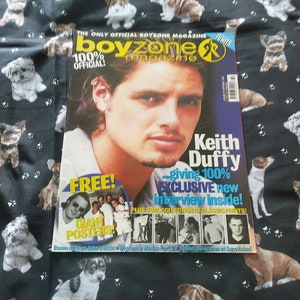 May include: A Boyzone magazine with Keith Duffy on the cover. The magazine has a blue and yellow design, with the text "100% Official!" and "Exclusive new interview inside!" The background is a black fabric with dog illustrations.