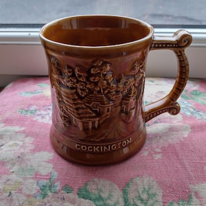 Peut inclure: Une chope en céramique marron avec un motif en relief détaillé de bâtiments et d'arbres. La chope a une anse décorative et le mot "COCKINGTON" est imprimé sur la base. La chope est posée sur une surface à motifs floraux roses.