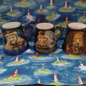 Puede incluir: Tres tazas de cerámica con retratos de hombres mayores. Las tazas tienen diferentes diseños de asa y combinaciones de colores, incluyendo azul, marrón y negro. La tela de fondo presenta un patrón náutico con faros y olas.