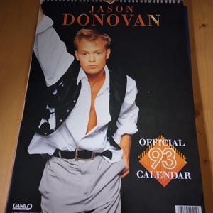 May include: A 1993 calendar featuring Jason Donovan. The calendar has a black background with the text "JASON DONOVAN" in orange and gold. The image shows Donovan in a white shirt, black vest, and grey trousers. The calendar also has the text "OFFICIAL 93 CALENDAR".