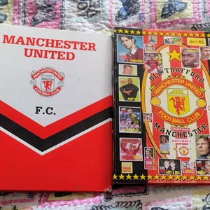 May include: Two vintage Manchester United football club items. One is a red, white, and black book with "MANCHESTER UNITED" and "F.C." printed on the cover. The other is a sticker album with player images and the club logo.