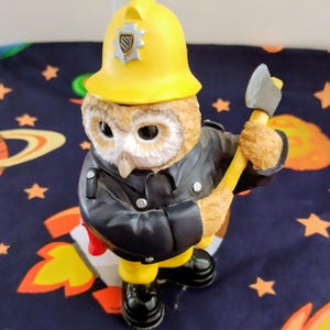 May include: A ceramic owl figurine in a firefighter's uniform, complete with a yellow helmet and axe. The owl is wearing a black jacket and yellow boots. The background is a blue fabric with star and planet patterns.