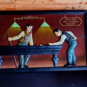 Pode incluir: Escultura em relevo emoldurada retratando duas figuras jogando bilhar. As figuras estão vestidas com trajes vintage, posicionadas em torno de uma mesa de bilhar escura. O fundo é vermelho escuro com duas lâmpadas verdes e uma placa que diz "OPEN DAILY NEW MEMBERS WELCOME."