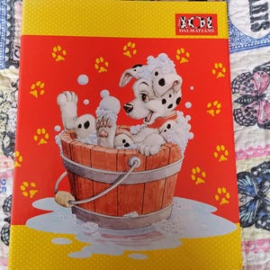 May include: A red and yellow notebook featuring a Dalmatian puppy in a wooden tub filled with bubbles. The cover has paw prints and the words "101 Dalmatians". The notebook has a yellow border.