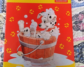 Peut inclure: Un cahier rouge et jaune avec un chiot dalmatien dans une baignoire en bois remplie de bulles. La couverture porte des empreintes de pattes et les mots "101 Dalmatians". Le cahier a une bordure jaune.
