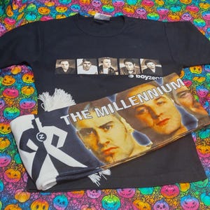 Puede incluir: Camiseta negra con una foto de la banda y la palabra "boyzone". Una bufanda con las palabras "THE MILLENNIUM" y caras de miembros de la banda está encima de la camiseta. También es visible una bufanda blanca y negra con un gráfico.