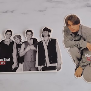 May include: Two die-cut stickers featuring black and white photographs of the boy band Take That. One sticker shows the five members standing together with the text Take That TV HITS. The second sticker shows a single member with the text Mark TV HITS.