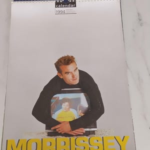 Puede incluir: Un calendario de pared de 1994 con una fotografía de Morrissey vistiendo una camisa negra de manga larga y abrazando un televisor. La pantalla muestra una escena con una persona en una camiseta amarilla. El nombre MORRISSEY está impreso en grandes letras amarillas en la parte inferior.