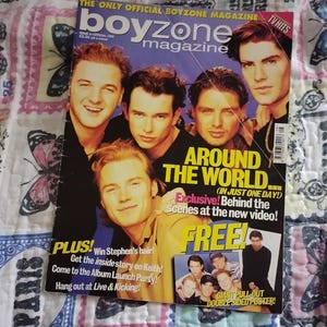 May include: A Boyzone magazine cover with five men on the front. The magazine has the text "AROUND THE WORLD" and "FREE!" The magazine's title is "THE ONLY OFFICIAL BOYZONE MAGAZINE". A smaller image of the band is in the bottom right corner. The magazine is on a patterned surface.