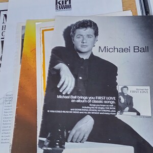 Pode incluir: Um anúncio impresso para o álbum First Love de Michael Ball, apresentando um retrato em preto e branco do artista com camisa escura, com texto listando títulos de músicas e o nome do álbum, sobre outros documentos de papel musicais.