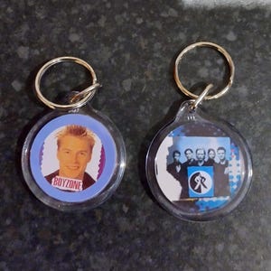 May include: Two clear acrylic keychains with silver-colored key rings. One keychain features a photo of a blonde man with the word "BOYZONE" below. The other keychain has a photo of a group of men.