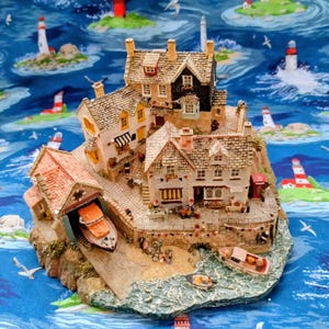 May include: A detailed miniature coastal village scene featuring houses and a boathouse. The buildings have textured roofs and are painted in various colors. A small boat is in the boathouse. The scene is set on a rocky island with a blue ocean background.