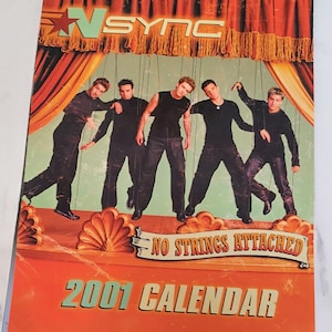 Könnte beinhalten: Ein N Sync Wandkalender für das Jahr 2001, der die Bandmitglieder in schwarzer Kleidung zeigt, die wie Marionetten mit angebrachten Fäden vor einer Bühnenkulisse mit orangefarbenen Vorhängen posieren. Der Text lautet N Sync, No Strings Attached und 2001 Calendar.