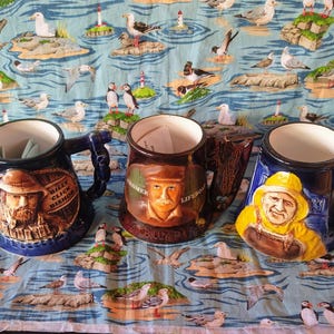 Peut inclure: Trois mugs en céramique avec des motifs nautiques. L'un est bleu marine avec un portrait d'homme chapeauté, un autre est marron avec un portrait d'homme en casquette, et le troisième est bleu avec un homme portant un ciré jaune.