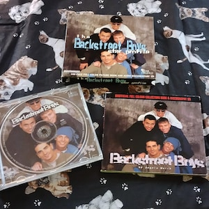 Könnte beinhalten: Ein CD und zwei Bücher mit den Backstreet Boys. Die CD befindet sich in einer transparenten Hülle, und die Bücher zeigen ein Gruppenfoto der Band. Die Bücher tragen die Titel "Backstreet Boys Star Profile" und "Official Full Color Collectors Book & Discography". Der Hintergrund ist ein schwarzer Stoff mit Hundeillustrationen.