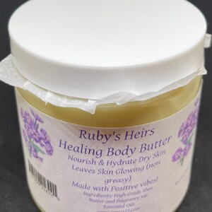 May include: A white jar with a white lid containing a light yellow body butter. The jar has a label that reads "Ruby's Heirs Healing Body Butter Nourish & Hydrate Dry Skin Leaves Skin Glowing (non greasy) Made with Positive vibes! Ingredients: High Grade Shea Butter and fragrance via Essential Oils www.tonya23.com"
