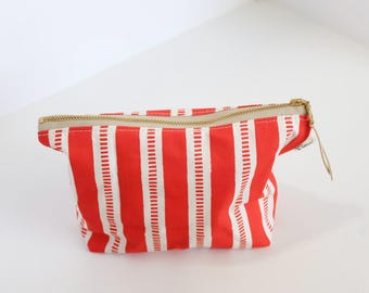 May include: A rectangular zippered pouch featuring a vibrant pattern of vertical red stripes alternating with white stripes that contain small, dashed red lines. The pouch is constructed with a gold-toned metal zipper closure along the top edge.