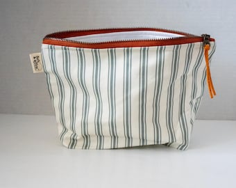 May include: A zippered pouch featuring a cream-colored fabric with vertical sage green stripes. The top edge has a rust-colored zipper with a matching leather pull tab. A small white fabric tag on the side displays the text fern & willow.