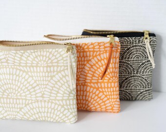 May include: Three rectangular fabric pouches with gold-tone metal zippers and leather pulls. The pouches feature a repeating fan-like pattern in beige, orange, and black. A small white tag on the beige pouch displays the text forth & arrow.