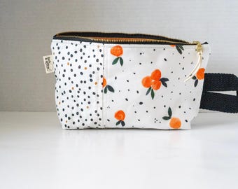May include: A white zippered pouch featuring a black polka dot pattern on the left side and an orange fruit print with green leaves on the right side. The bag includes a black fabric side handle, a gold-tone zipper, and a small white brand tag.