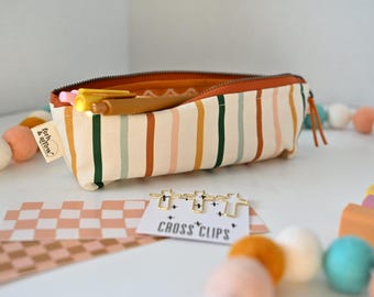 May include: A rectangular fabric pencil case with vertical stripes in shades of cream, mustard yellow, dark green, and terracotta. The case features a brown zipper and a small tag labeled fern & arrow. Inside are two pens and gold cross clips.