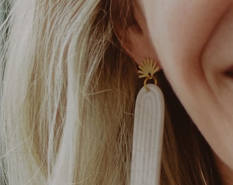 May include: A dangle earring featuring a gold-toned metal sunburst stud connected to a long, arched, off-white clay pendant. The pendant has a subtle speckled texture and three vertical embossed lines following the curve of the arch shape.
