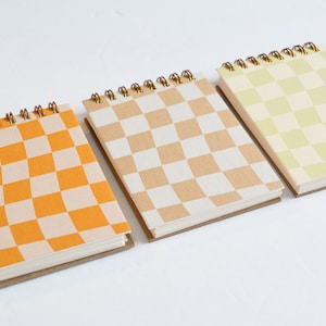 May include: Three spiral-bound notebooks with checkered covers in orange, tan, and light green. Each notebook has a brown cardboard base and gold-colored spiral binding. The notebooks are arranged on a white surface.