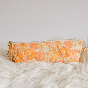 May include: A rectangular pencil case with a floral pattern in shades of orange, yellow, and cream. The case has a zipper closure with a gold-colored pull and a small orange wrist strap. The case is approximately 8 inches long.
