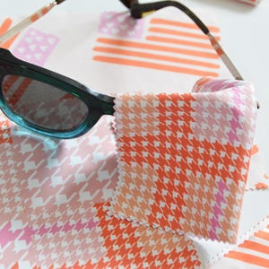 May include: A pair of teal-framed sunglasses rests on patterned cleaning cloths. The cloths feature a houndstooth design in shades of pink, orange, and white. Additional cloths with a striped pattern are also visible.