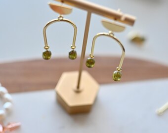 May include: A pair of gold-toned earrings featuring a semi-circular top piece and an inverted U-shaped arch. Each end of the arch is adorned with a small, translucent olive-green bead. The earrings are displayed on a gold-toned metal stand.