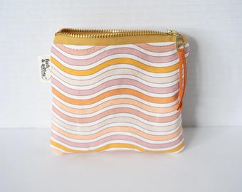 May include: A small rectangular fabric pouch featuring a repeating pattern of horizontal wavy lines in shades of pink, orange, yellow, and cream. The pouch has a gold-toned metal zipper with a thin orange leather pull and a white side tag reading fern & arrow.