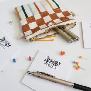 May include: A brown and white checkered fabric pouch with a zipper closure. The pouch is filled with two silver pens and has a colorful striped fabric lining. The pouch is sitting on a white surface with several colorful push pins scattered around it. There are also two white notepads with black text that reads "Mickey & Co."