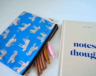 May include: A bright blue zippered pouch featuring a repeating pattern of white and black cheetahs, with several pens tucked inside. Beside it lies a beige notebook with the text "NICE IDEAS" and "notes & thoughts" printed in blue.