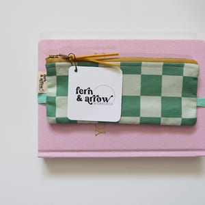 May include: A green and white checkered pencil case with a yellow zipper. The pencil case is sitting on top of a pink notebook. A white tag with the text "fern & arrow workshop" is attached to the zipper.