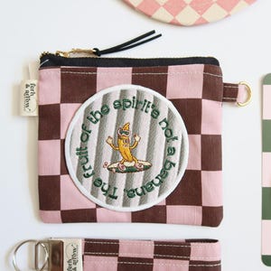 May include: A pink and brown checkered zippered pouch with a round embroidered patch that says "The fruit of the spirit's not a banana". The patch features a cartoon banana wearing sunglasses and riding a surfboard.