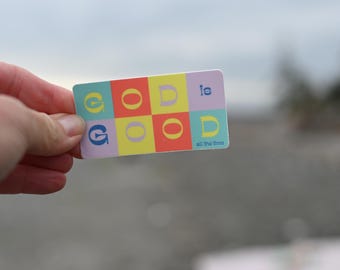 May include: A rectangular sticker with the words "GOD is GOOD all the time" in a colorful, blocky design. The letters are in various colors, including teal, yellow, red, and purple. The sticker is held against a blurred background.